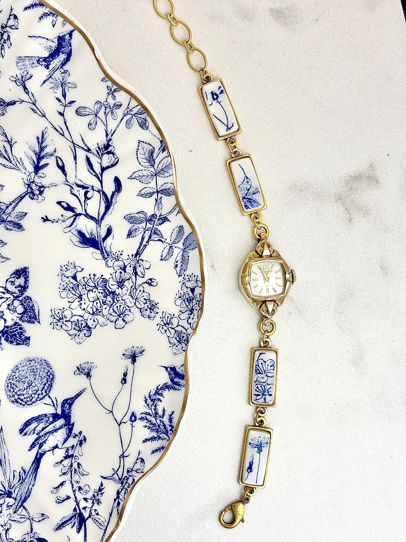 May include: A gold-tone link bracelet watch featuring rectangular ceramic tiles with blue and white floral and hummingbird patterns. The watch has a square face with gold hands and markers, connected by gold metal links and a lobster clasp closure.