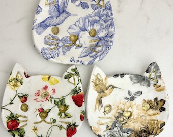 May include: Three cat-shaped ceramic trinket dishes. The top dish features a blue and white floral pattern with a hummingbird. The bottom left dish displays strawberries and flowers. The bottom right dish shows a hummingbird and beige floral motifs.
