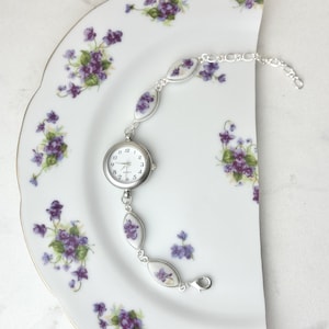 May include: A silver watch bracelet with oval links and a round watch face. The links and watch face feature purple floral designs. The watch is displayed on a white plate decorated with purple floral patterns.