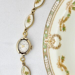 Puede incluir: Un reloj vintage dorado con esfera blanca y la marca "UGANT". El reloj est&aacute; conectado a una pulsera con eslabones ovalados blancos con dise&ntilde;os florales. Tambi&eacute;n se ve una porci&oacute;n de un plato color crema con dise&ntilde;os florales verdes y dorados.