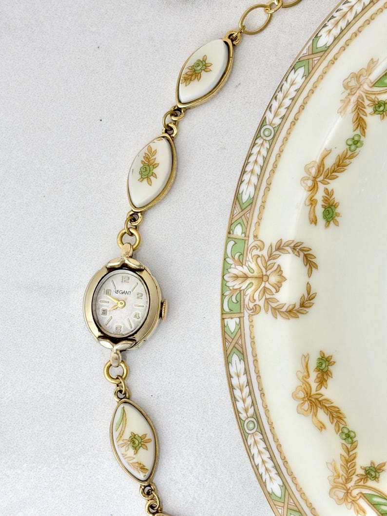 Puede incluir: Un reloj vintage dorado con esfera blanca y la marca "UGANT". El reloj est&aacute; conectado a una pulsera con eslabones ovalados blancos con dise&ntilde;os florales. Tambi&eacute;n se ve una porci&oacute;n de un plato color crema con dise&ntilde;os florales verdes y dorados.