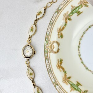 Puede incluir: Un brazalete de reloj dorado vintage con una esfera ovalada y detalles florales blancos y verdes decorativos. El brazalete est&aacute; junto a un plato decorativo con dise&ntilde;os florales verdes y dorados.