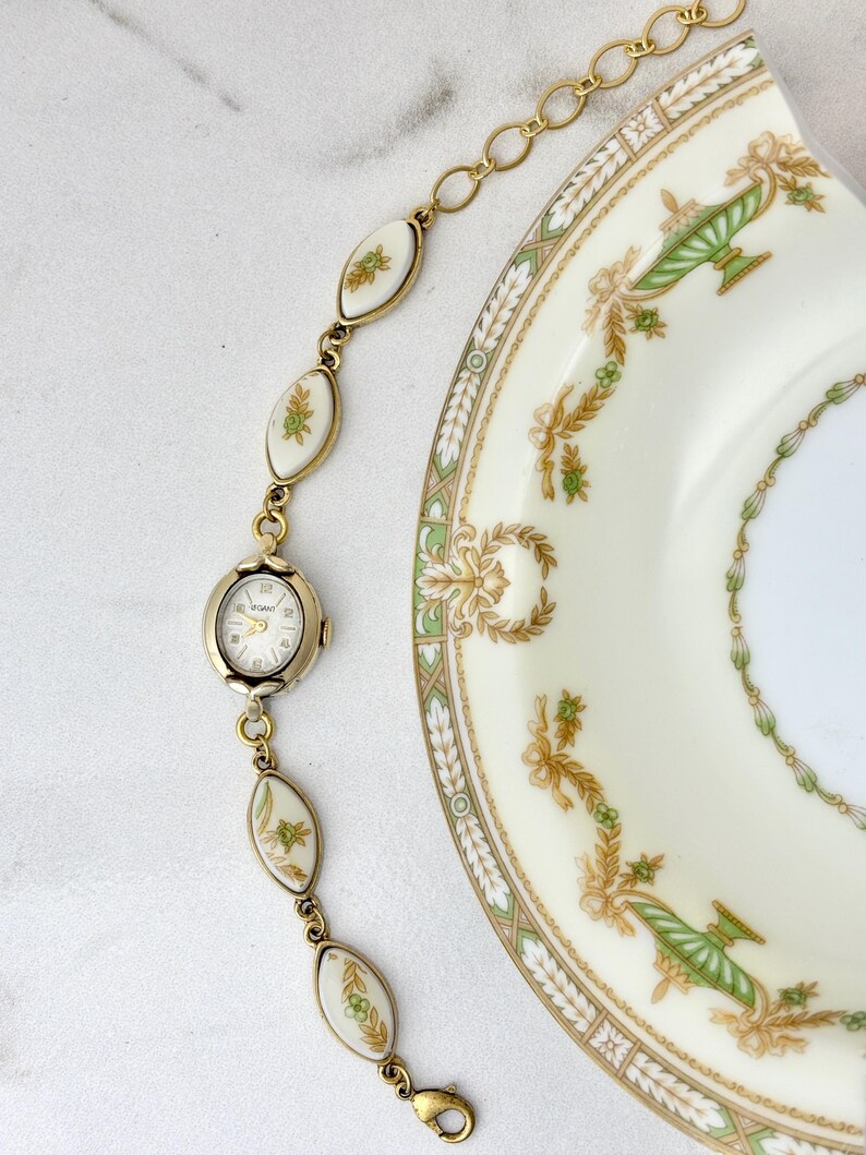 Puede incluir: Un brazalete de reloj dorado vintage con una esfera ovalada y detalles florales blancos y verdes decorativos. El brazalete est&aacute; junto a un plato decorativo con dise&ntilde;os florales verdes y dorados.