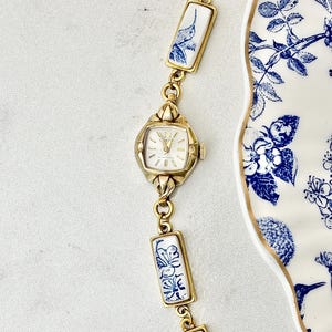 May include: A gold-tone wristwatch with a white dial and gold hands, featuring a bracelet composed of rectangular white ceramic links decorated with blue floral and hummingbird patterns, positioned next to a matching blue and white floral plate.