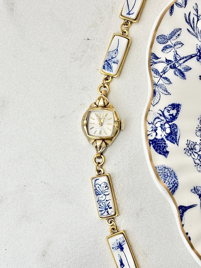 May include: A gold-tone wristwatch with a white dial and gold hands, featuring a bracelet composed of rectangular white ceramic links decorated with blue floral and hummingbird patterns, positioned next to a matching blue and white floral plate.