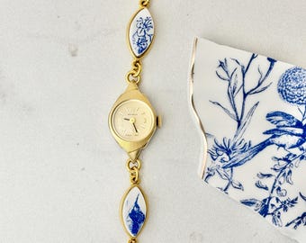 Op de afbeelding: Een goudkleurig polshorloge met een ronde wijzerplaat en een schakelarmband met witte, marquisevormige keramische inzetstukken voorzien van blauwe bloemmotieven. De wijzerplaat toont de merknaam Benrus en goudkleurige wijzers.