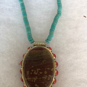 May include: A turquoise beaded necklace with an oval pendant. The pendant features a brown and tan stone set in a gold-colored metal frame, accented with red beads. The necklace is a unique piece of jewelry.