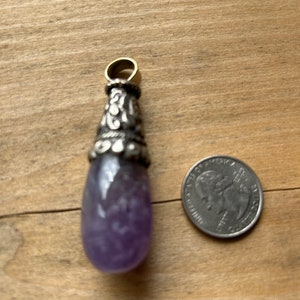 May include: A teardrop-shaped pendant with a purple gemstone and silver-colored metal accents. The pendant has a gold-colored loop at the top. A U.S. quarter is next to the pendant for size comparison.