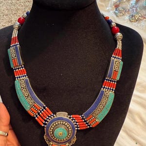 May include: A colorful necklace with a central pendant and curved sections. The necklace features turquoise, red, blue, and silver-toned beads and accents. The pendant has a circular design with a turquoise center.