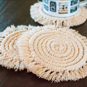May include: Two beige crocheted coasters with fringe, stacked on top of each other. The coasters are round and have a spiral pattern.
