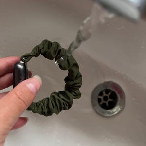 May include: A green fabric watch band is being rinsed under running water in a white sink.
