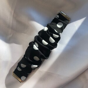 May include: Black scrunchie with white polka dots and gold-coloured metal clasps. The fabric is gathered, creating a textured look. A hair accessory.
