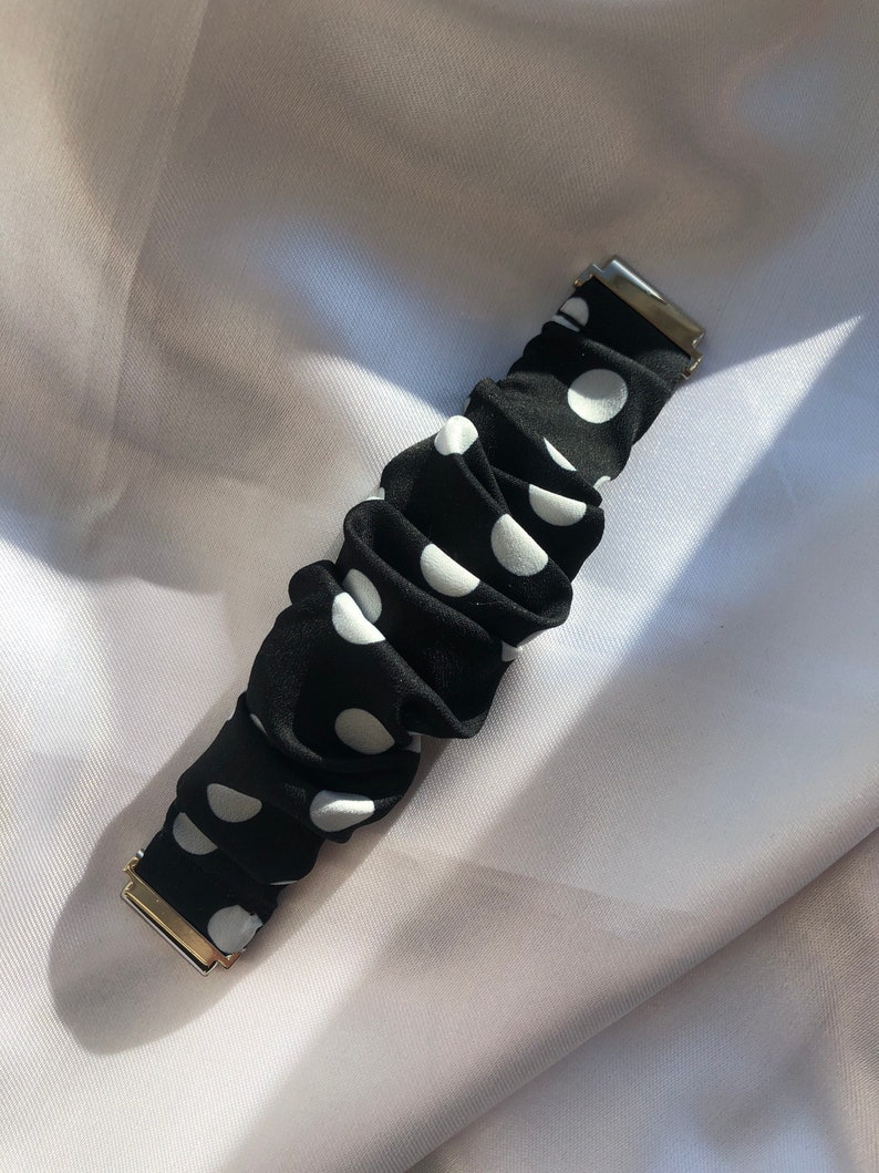 May include: Black scrunchie with white polka dots and gold-coloured metal clasps. The fabric is gathered, creating a textured look. A hair accessory.
