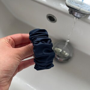 May include: A dark blue fabric hair scrunchie with a gathered, ruffled design. The scrunchie is being held in a hand over a white porcelain sink.