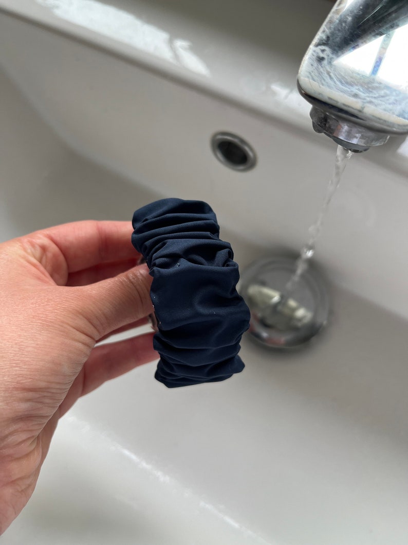 May include: A dark blue fabric hair scrunchie with a gathered, ruffled design. The scrunchie is being held in a hand over a white porcelain sink.