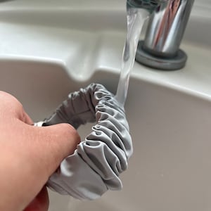 Puede incluir: Un scrunchie de tela gris se enjuaga bajo un chorro de agua en un lavabo blanco.