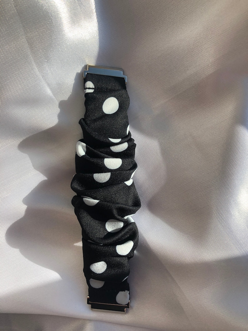 May include: Black and white polka dot watch strap with silver-coloured metal clasps. The strap is made of a ruched fabric with large white polka dots on a black background. The watch strap is shown against a white background.