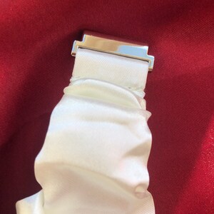 May include: Close-up of a white satin scrunchie with a silver-colored metal clasp. The scrunchie is gathered and has a soft, flowing texture. The background is a deep red fabric.