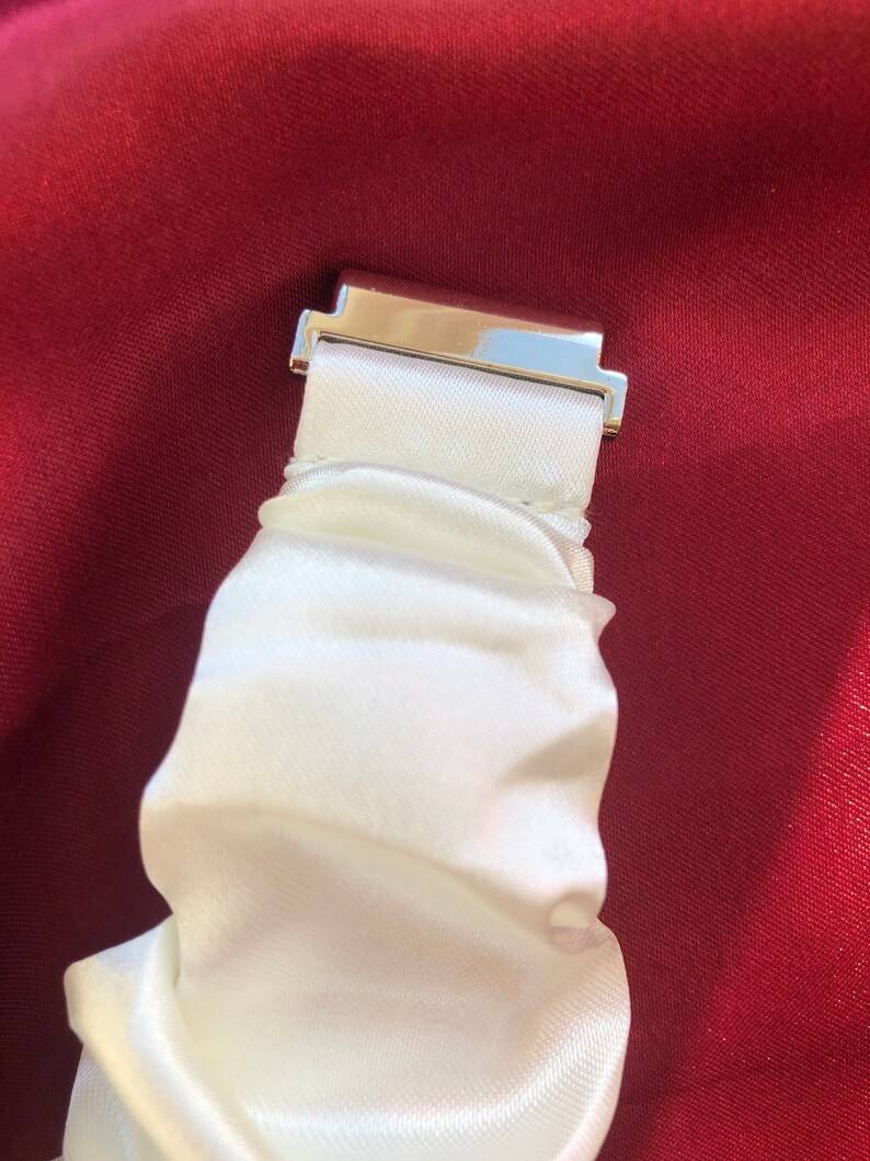 May include: Close-up of a white satin scrunchie with a silver-colored metal clasp. The scrunchie is gathered and has a soft, flowing texture. The background is a deep red fabric.