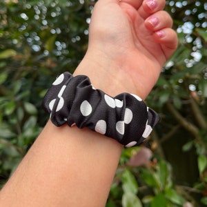 May include: A black scrunchie with white polka dots is wrapped around a wrist. The scrunchie is made of a silky material and is gathered, creating a textured appearance. The background is a blurred view of green foliage.