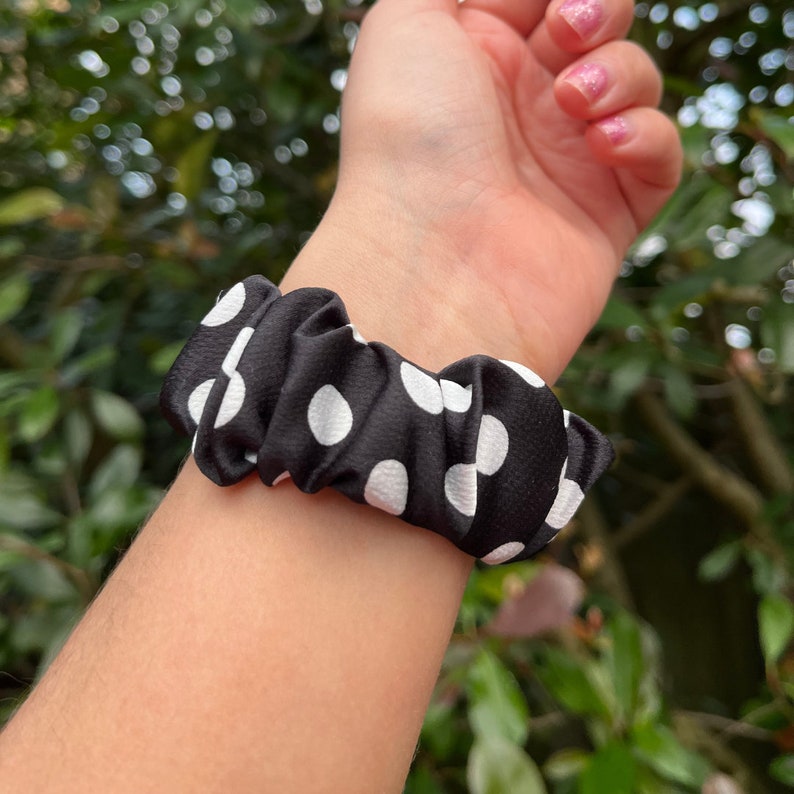 May include: A black scrunchie with white polka dots is wrapped around a wrist. The scrunchie is made of a silky material and is gathered, creating a textured appearance. The background is a blurred view of green foliage.