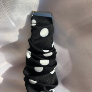 May include: Black scrunchie with white polka dots and a silver metal clasp. The fabric is gathered, creating a textured appearance. The polka dots are evenly spaced across the black material.