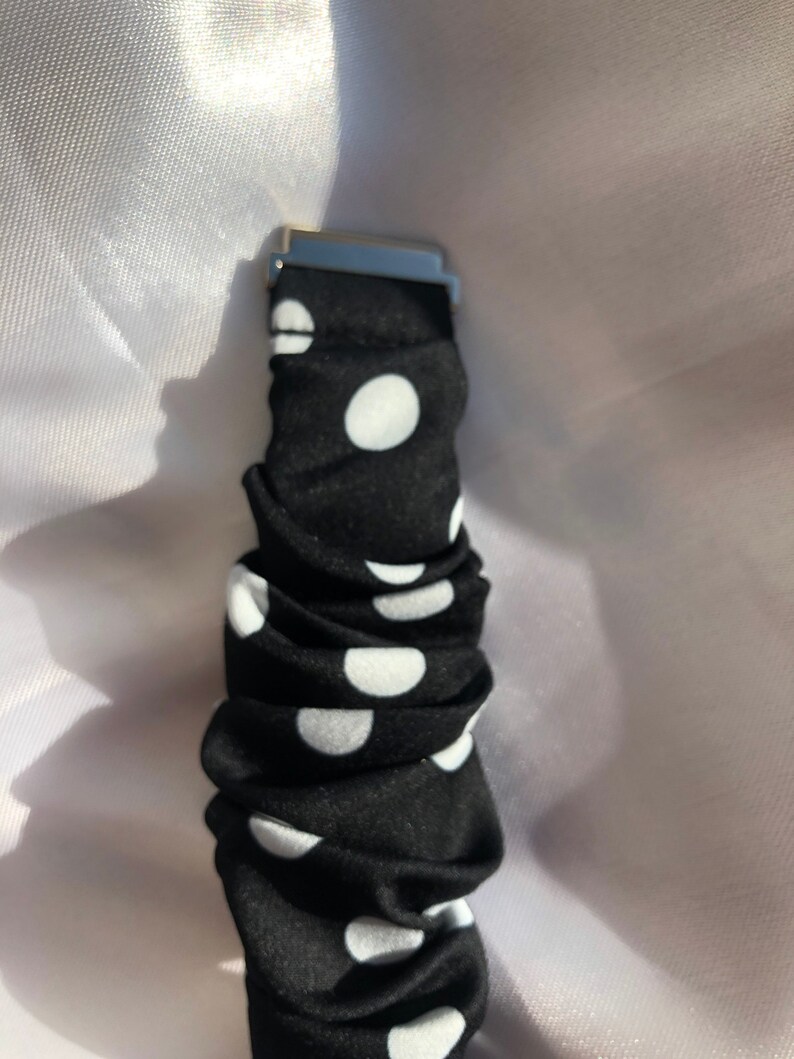 May include: Black scrunchie with white polka dots and a silver metal clasp. The fabric is gathered, creating a textured appearance. The polka dots are evenly spaced across the black material.