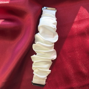 May include: A white, ruffled fabric watch band with silver-colored metal clasps. The band is made of a soft, silky material and is gathered in a series of horizontal folds. The background is a deep red fabric.