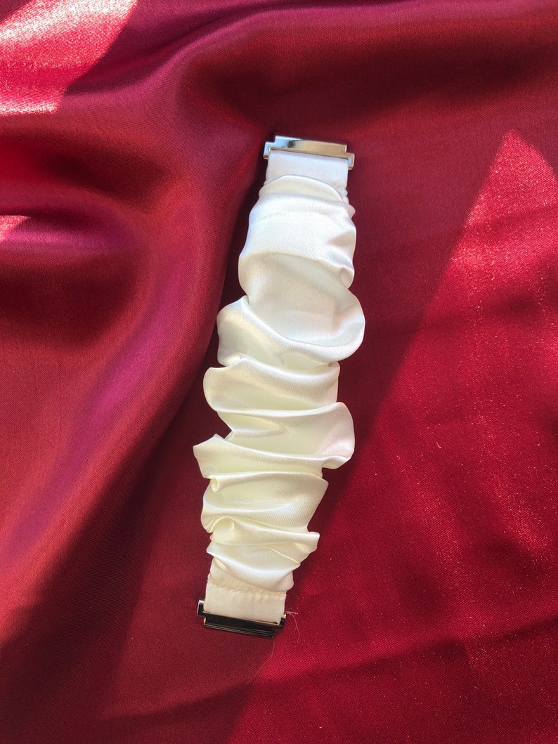 May include: A white, ruffled fabric watch band with silver-colored metal clasps. The band is made of a soft, silky material and is gathered in a series of horizontal folds. The background is a deep red fabric.