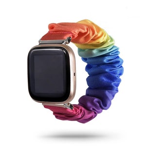 Puede incluir: Un reloj inteligente con una caja dorada y una correa de scrunchie de color arcoíris. La correa hace la transición por los colores del arcoíris, del rojo al morado. La esfera del reloj es negra.