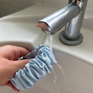 Könnte beinhalten: Ein hellblauer Stoff-Haargummi wird unter einem silbernen Wasserhahn in einem weißen Porzellanwaschbecken gespült.