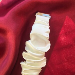 May include: A white, ruffled fabric wristband with a silver-colored clasp. The wristband is made of a silky material and is gathered in a series of folds. The background is a deep red fabric.