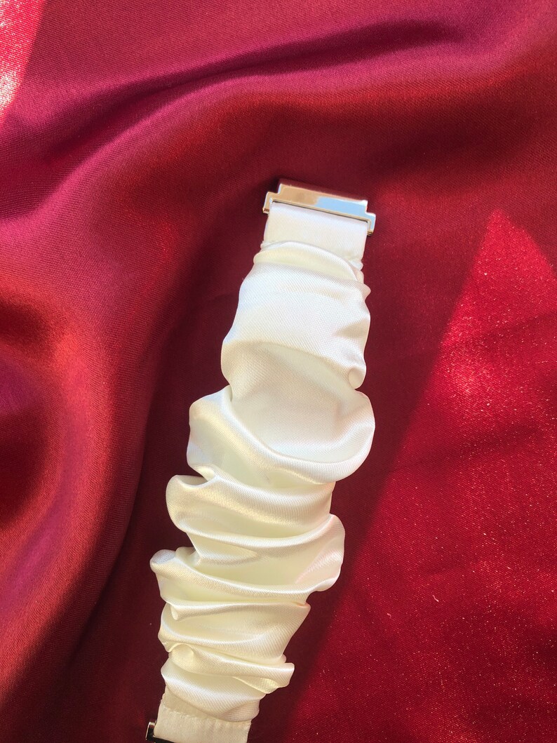 May include: A white, ruffled fabric wristband with a silver-colored clasp. The wristband is made of a silky material and is gathered in a series of folds. The background is a deep red fabric.