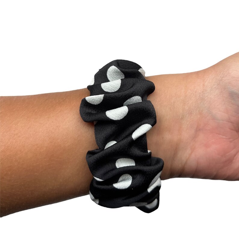 May include: Black scrunchie with white polka dots. The fabric is gathered and wrapped around the wrist. A hair accessory.