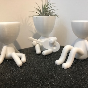 May include: Three white ceramic planters shaped like people, one playing a guitar, sitting on a black surface.