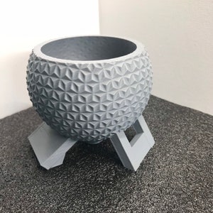 May include: A gray, geometrically patterned planter with a stand. The planter is shaped like a sphere and has a textured surface. The stand is made of the same material as the planter and has three legs.