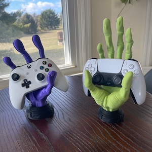May include: Two 3D printed hands holding video game controllers. The hand on the left is purple and holding a white Xbox controller. The hand on the right is green and holding a white Playstation 5 controller.