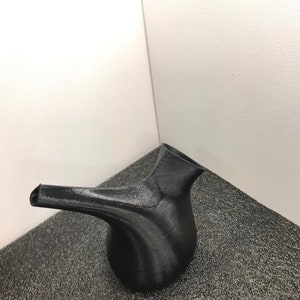 May include: A black ceramic teapot with a curved spout and a rounded body. The teapot is sitting on a textured black surface.