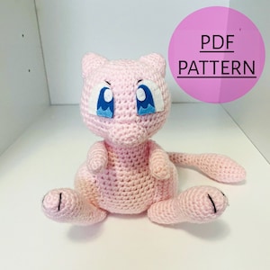 May include: A handmade pink crochet Mew plushie with large blue eyes and a long tail. The plushie is sitting with its legs extended. A pink circle with the text "PDF PATTERN" is in the background.