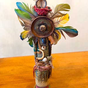 Puede incluir: Una escultura decorativa vertical con una columna cilíndrica central envuelta en tela texturizada verde y beige, adornada con un medallón de madera circular, plumas coloridas, conchas pequeñas, cordel y una base sobre un soporte cuadrado.