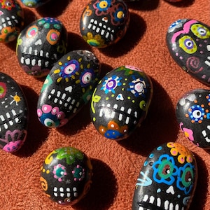 May include: A collection of hand-painted black stones, each adorned with colorful sugar skull designs. The skulls feature white teeth, floral patterns, and vibrant accents in pink, blue, yellow, and green. The stones vary in size and shape.