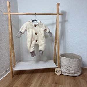 May include: A cream-colored baby jumpsuit with a hood and brown buttons hangs on a wooden clothing rack. The rack has a shelf and is next to a woven basket with a rainbow design. A wooden disc with the text "NOEM" is also present.