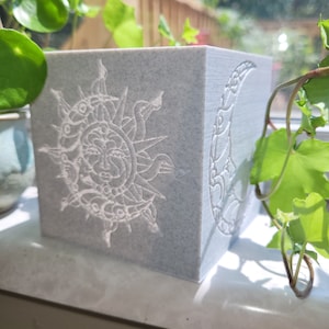 May include: A white ceramic cube with a sun and moon design. The sun has a face and the moon is a crescent shape. The cube is sitting on a windowsill.