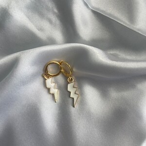 May include: A pair of gold hoop earrings with white lightning bolt charms. The earrings are on a white satin background.
