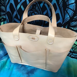 May include: A light beige leather tote bag featuring two top handles, two exterior front pockets, and a central embossed logo patch. The bag is constructed with visible white stitching along the seams and handles, showcasing a natural finish.