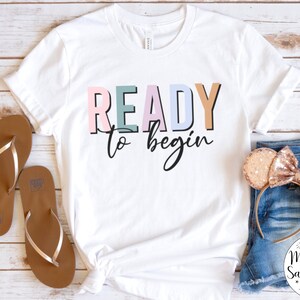 K&ouml;nnte beinhalten: Wei&szlig;es T-Shirt mit dem Text "READY to begin" in bunten Buchstaben. Das Shirt liegt flach auf einer Holzoberfl&auml;che mit einem Paar braunen Flip-Flops und einem Paar goldenen Glitzer-M&auml;useohren.