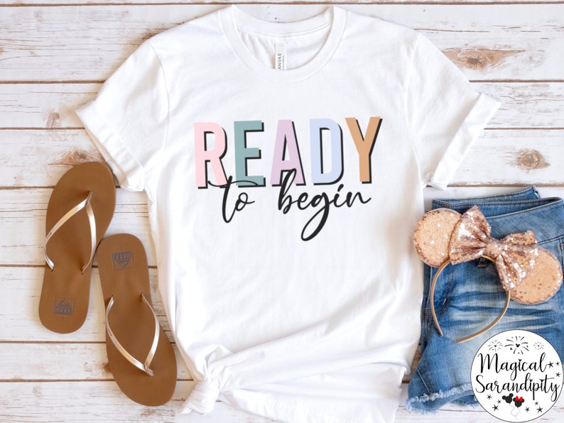 K&ouml;nnte beinhalten: Wei&szlig;es T-Shirt mit dem Text "READY to begin" in bunten Buchstaben. Das Shirt liegt flach auf einer Holzoberfl&auml;che mit einem Paar braunen Flip-Flops und einem Paar goldenen Glitzer-M&auml;useohren.
