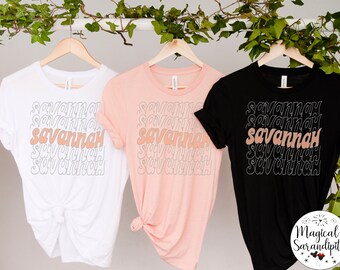 May include: Three t-shirts with the word "Savannah" repeated in a stacked pattern. The shirts are white, pink, and black. The word "Savannah" is in a peachy-orange color.