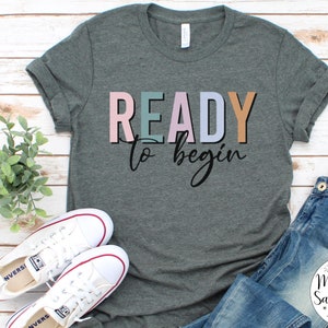 K&ouml;nnte beinhalten: Ein heidefarbenes T-Shirt mit dem Text "READY to begin" in bunten Blockbuchstaben. Das Shirt liegt flach auf einer wei&szlig;en Holzoberfl&auml;che mit einem Paar wei&szlig;en Turnschuhen und einer blauen Jeans.