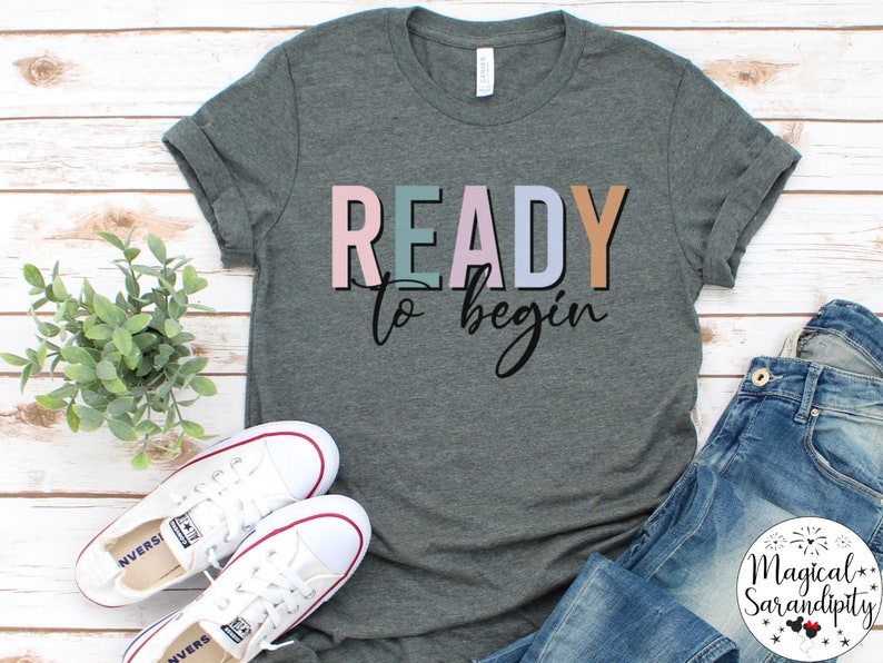 K&ouml;nnte beinhalten: Ein heidefarbenes T-Shirt mit dem Text "READY to begin" in bunten Blockbuchstaben. Das Shirt liegt flach auf einer wei&szlig;en Holzoberfl&auml;che mit einem Paar wei&szlig;en Turnschuhen und einer blauen Jeans.